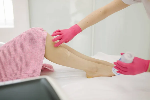 Young woman beautician in mask applies contact gel on the leg of a woman for laser hair removal in medical clinic. Close up. Pink glowes — Stock Photo, Image