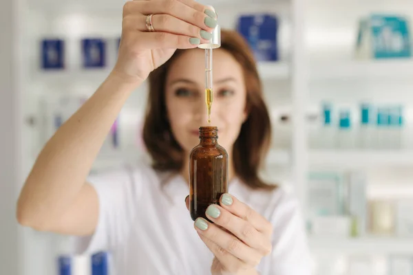 Una doctora cosmetóloga con una bata blanca sostiene una botella de aceite de argán enriquecido en las manos. Mujer con pipeta. Producto de cuidado facial en salón de belleza. Sala de Cosmetología — Foto de Stock