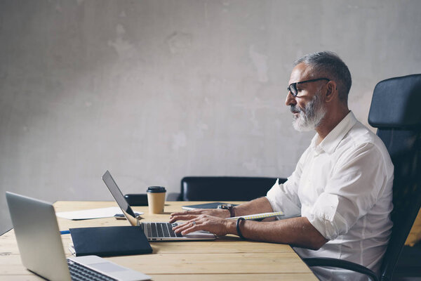 Positive Adult businessman using mobile laptop computer while sitting at wooden table at modern coworking place. .