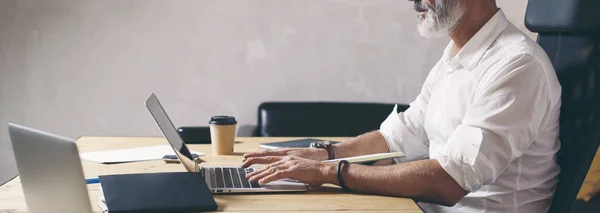 Empresário adulto atraente e confidente usando o computador portátil móvel enquanto trabalhava na mesa de madeira no escritório de coworking moderno. Ampla . — Fotografia de Stock