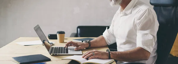 Pozitivní vousatý podnikatel pomocí mobilní přenosný počítač zatímco sedí u dřevěného stolu na místě moderní coworkingu. Široké — Stock fotografie