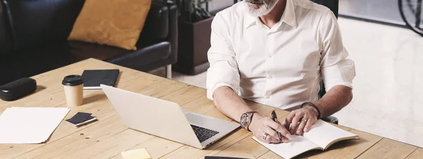 Atrakcyjny i poufnych biznesmen dorosłych za pomocą mobilnego laptopa i dokonywanie notatek podczas pracy przy stole drewniane nowoczesne coworking urzędzie. Szeroki — Zdjęcie stockowe