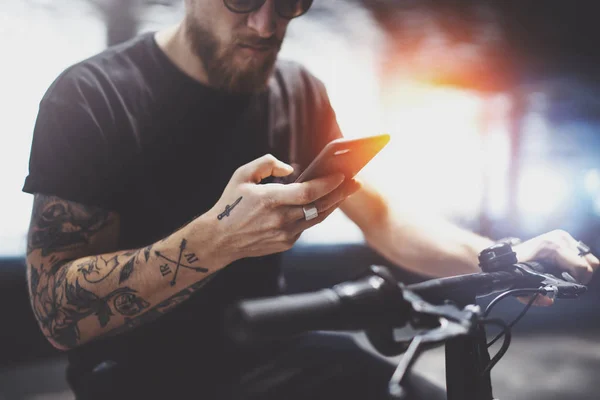 Homme tatoué barbu dans des lunettes de soleil en utilisant un téléphone mobile pour envoyer un message texte après avoir roulé en scooter électrique dans la ville . — Photo