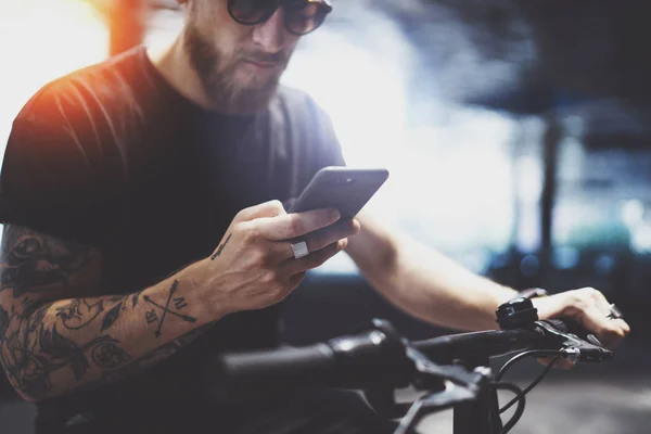 Hombre tatuado barbudo en gafas de sol usando teléfono móvil para enviar mensaje de texto después de montar en scooter eléctrico en la ciudad . — Foto de Stock