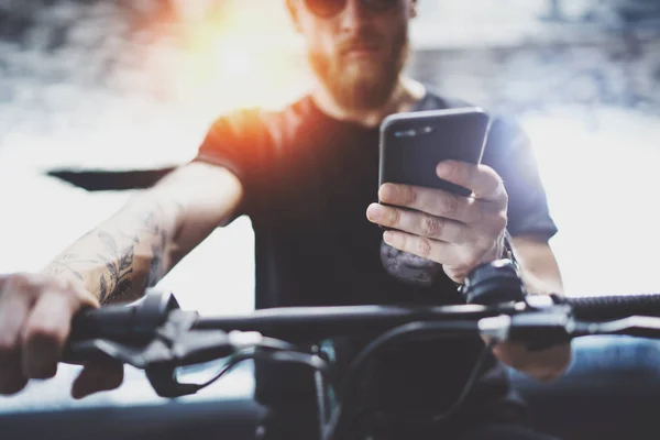 Hipster musculoso barbudo tatuado en gafas de sol usando smartphone después de montar en scooter eléctrico en la ciudad. Transporte innovador . — Foto de Stock