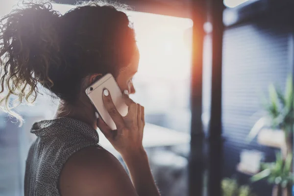 Mujer joven guapo hablando con amigos a través de un teléfono inteligente moderno mientras pasa su tiempo en la cafetería urbana moderna . — Foto de Stock