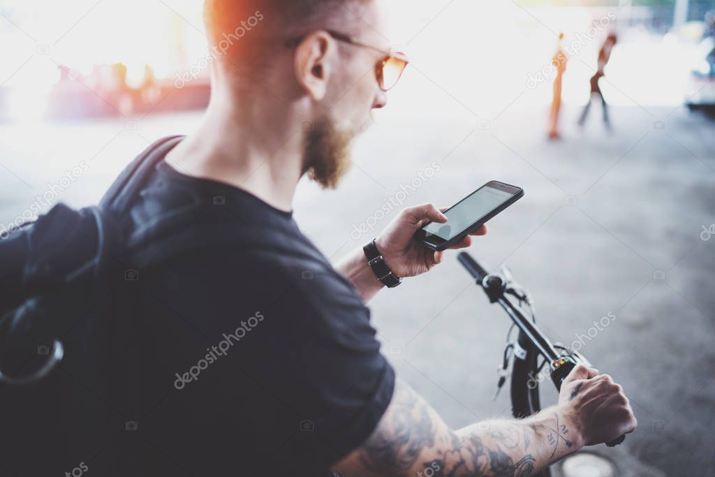 Bearded muscular tattooed hipster in sunglasses using smartphone after riding by electric scooter in the city. Innovative transport.
