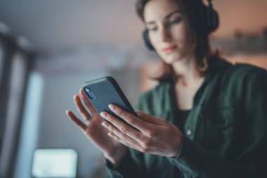 Modern evde rahatlayan, günlük kıyafetler giyen, akıllı telefondan müzik çalan ve kablosuz kulaklıkla ses kitapları dinleyen genç bir kız..