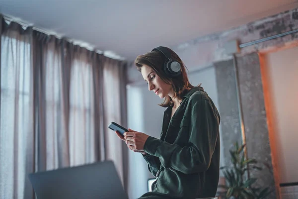 Młoda, przystojna dziewczyna relaksująca się we współczesnym domu, nosząca luźne ubrania, grająca muzykę na smartfonie i słuchająca książek audio w słuchawkach bezprzewodowych.Niewyraźne tło. — Zdjęcie stockowe