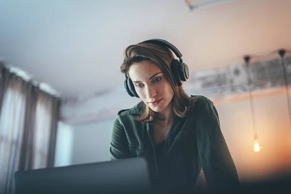 Menina bonita jovem relaxante em casa moderna, vestindo roupas casuais, tocando música no smartphone e ouvindo livros de áudio em fones de ouvido sem fio.Fundo desfocado . — Fotografia de Stock