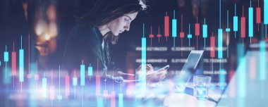 İş kadını gece ofisinde dizüstü bilgisayarın önünde oturuyor ve cep telefonunu kullanıyor. Kırmızı ve yeşil mum çubuğu grafik ve hisse senedi ticareti bilgisayar ekran arka plan. Çifte pozlama. Geniş.