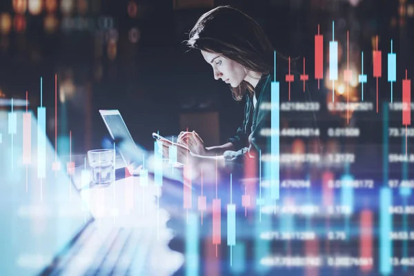 Geschäftsfrau sitzt im Nachtbüro vor Laptop-Computer mit Finanzgrafiken und bedient ihr Smartphone. roter und grüner Leuchter Chart und Aktienhandel auf Hintergrund. Doppelbelastung. — Stockfoto