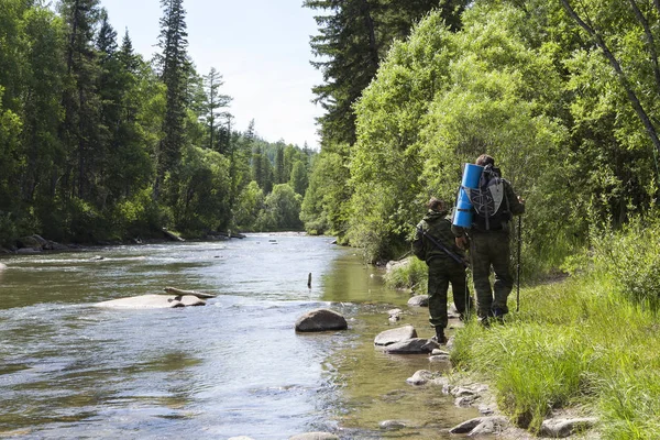 The forester and the inspector with a backpack, sleeping mat behind his back and a rifle in the trek across the mountain river, flowing among the stones in the forest