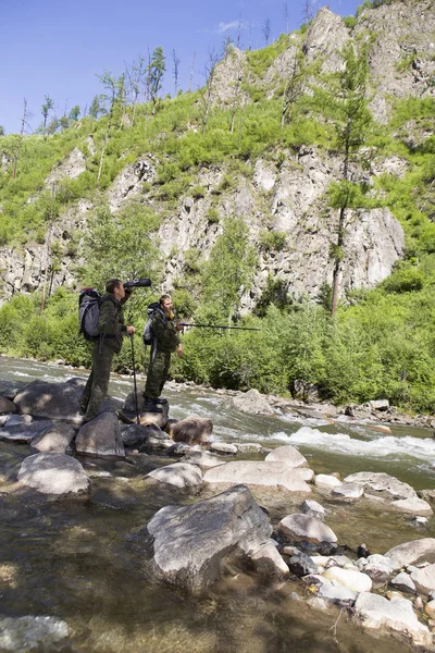 The forester and the inspector with a backpack, sleeping mat behind his back and a rifle in the trek across the mountain river, flowing among the stones in the forest