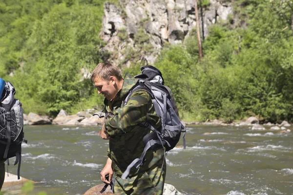 The forester and the inspector with a backpack, sleeping mat behind his back and a rifle in the trek across the mountain river, flowing among the stones in the forest