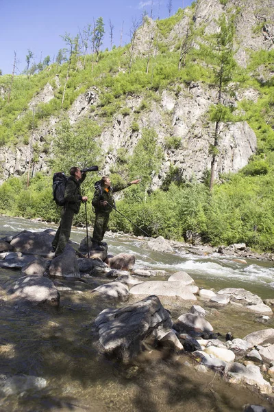 The forester and the inspector with a backpack, sleeping mat behind his back and a rifle in the trek across the mountain river, flowing among the stones in the forest