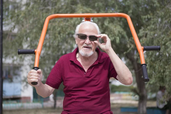 Attractive senior man trains on sporting equipment in a city in the open air. The concept of a healthy lifestyle and accessibility of sports training for every person. Available sports equipment in a public place.