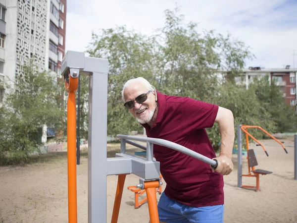 Attractive senior man trains on sporting equipment in a city in the open air. The concept of a healthy lifestyle and accessibility of sports training for every person. Available sports equipment in a public place.