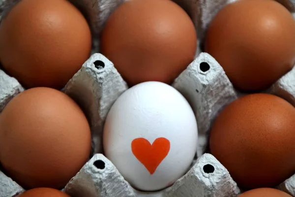 View from above of a white egg with a red heart painted on it, surrounded by five brown eggs in a egg box