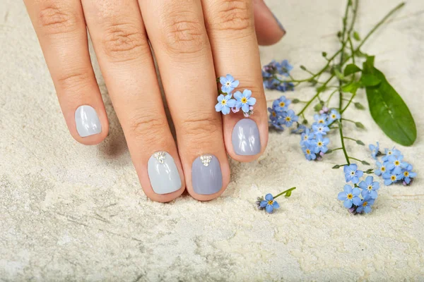 Hand with short manicured nails colored with gray nail polish and forget me not flowers