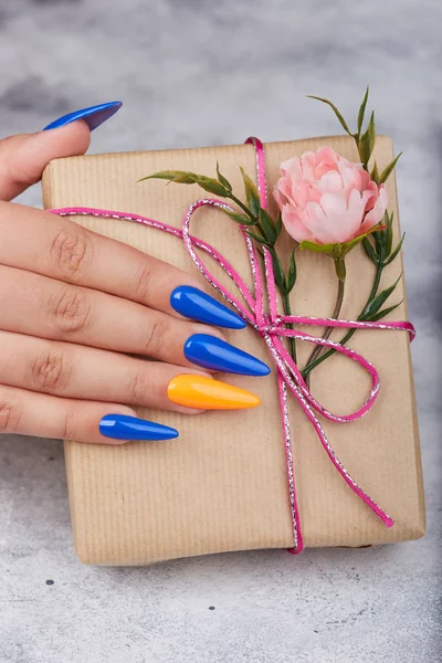 弓とピンクの花とギフトボックスを保持する青とオレンジのマニキュアで着色された人工手入れ爪と手 — ストック写真