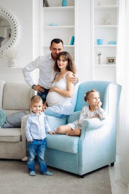 Üçüncü çocuklarını bekleyen büyük bir aile. Hamile bir kadın ve iki büyük sarışın çocuğu olan bir baba mavi bir sandalyede oturuyorlar. Mutlu aile hayatı. Aile fotoğrafı. Baba annesine sarılıyor.