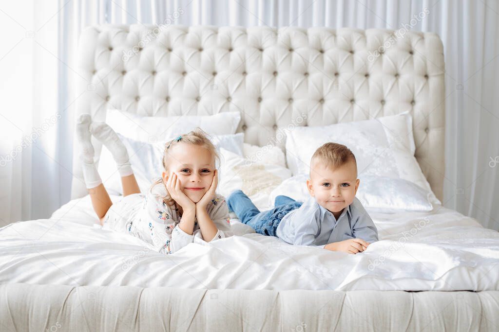 small children, a blond brother and sister lie on the bed of a white bed, smiling and looking at the camera