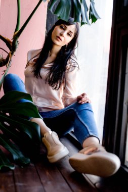 Genç Asyalı esmer, evin penceresinde tek başına oturmuş kameraya bakıyor. Kotlu ve pembe bluzlu kız. .