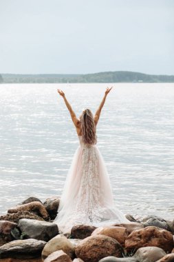 Saçı açık pembe bir gelinlik içindeki güzellik denize bakan bir kayanın üzerinde duruyor. Ellerini havaya kaldırdı..