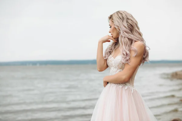 Belleza joven en la playa con vistas al mar. Retrato sensual de una novia sexy con el pelo suelto en un vestido rosa suave — Foto de Stock