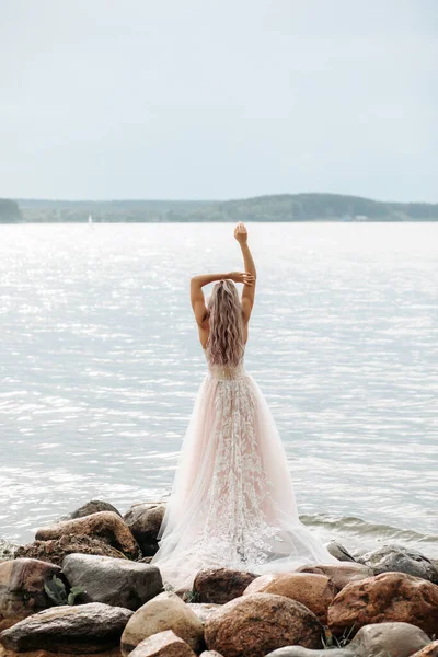 Egy menyasszony egy puha rózsaszín esküvői ruhában lehajtott hajjal áll egy sziklán, kilátással a tengerre.. — Stock Fotó
