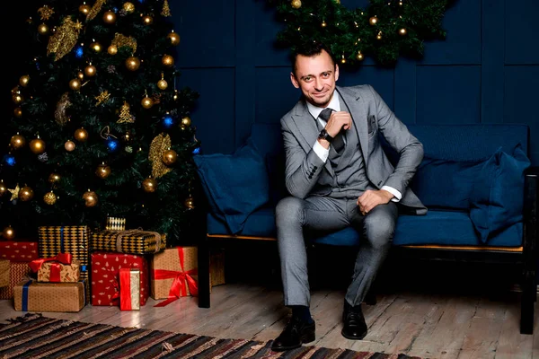 Yılbaşı gecesi ofiste gri takım elbiseli bir iş adamı. Kırmızı bir Noel ağacı ve hediye kutularının yanındaki mavi bir kanepede oturmak. Noel gecesini bekliyorum. Şenlik konsepti..