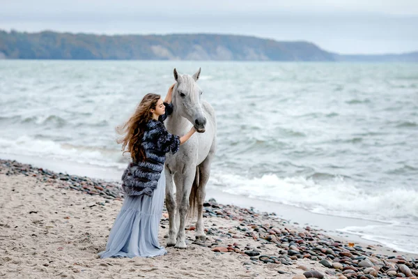 Una hermosa joven está hablando con un caballo blanco en el océano. caminando con un caballo en tiempo nublado en la playa. — Foto de Stock