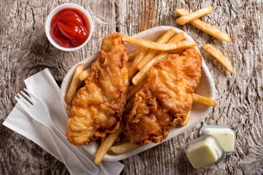 Lezzetli balık ve cips konteyner ile ketçap, tartar sosu, kağıt peçete ve rustik ahşap masa üstüne plastik çatal dışarı almak.