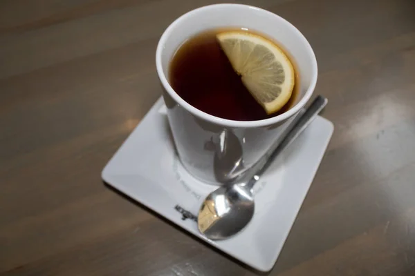 White porcelain cup with black hot tea and lemon, white porcelain saucer, a teaspoon on a brown table.