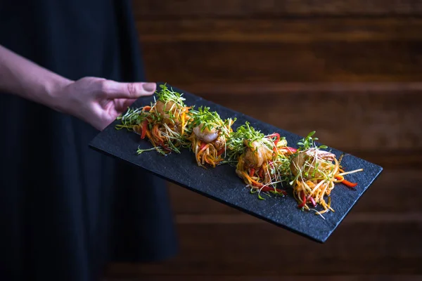 human hand holding seafood snack with vegetables on skewers served on slate