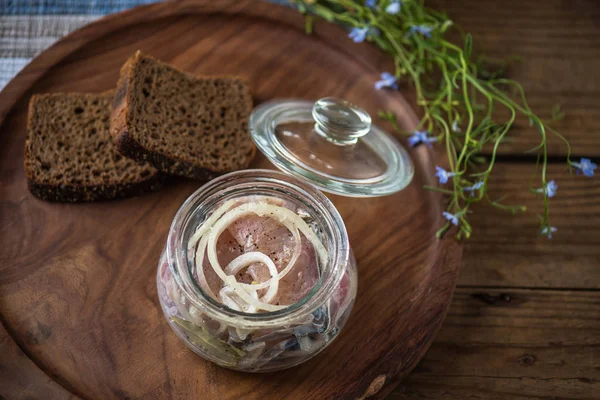 Pickled fish in glass jar with onion on wooden tray with slices of bread