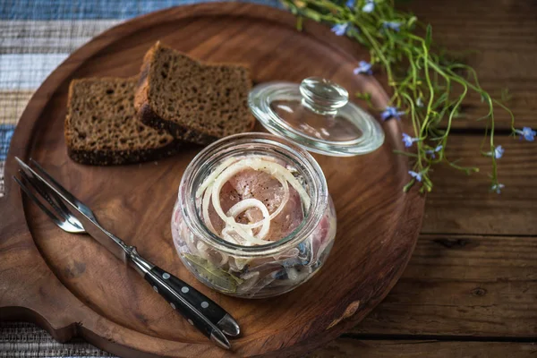 Pickled fish in glass jar with onion on wooden tray with slices of bread