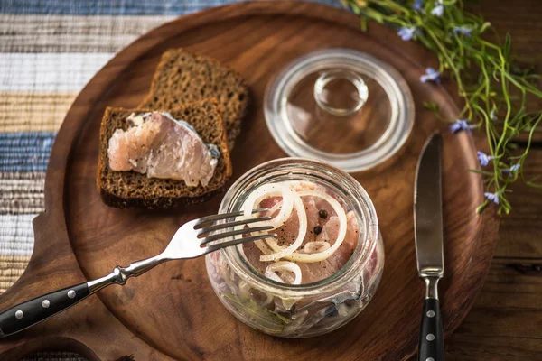 Pickled fish in glass jar with onion on wooden tray with slices of bread