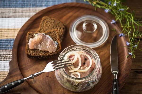 Pickled fish in glass jar with onion on wooden tray with slices of bread