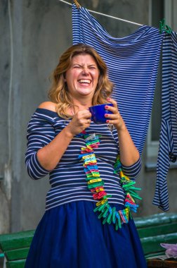 Bir bardak içkisi olan genç bir kadın çamaşırların yanında duruyor dışarıda, evin önünde, kırsal kesimde.