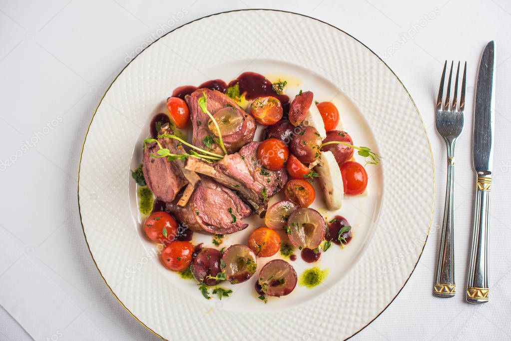 top view of delicious meat roasted with plums and tomatoes on white plate and fork with knife on table 