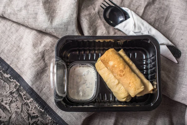 Rolled stuffed pancakes in plastic container with sauce on grey tablecloth
