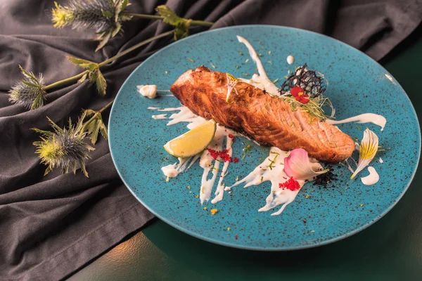 Fried piece of salmon with sauce and edible flowers on blue plate on wooden table