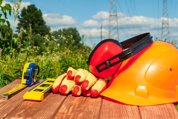 Capacete de proteção, fones de ouvido, luvas em uma mesa de madeira, contra o fundo da natureza e postes de alta tensão .. — Fotografia de Stock