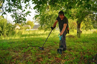 Yeşillik dolu canlı bir bahçede bir insan metal dedektörü kullanıyor. Öğleden sonra güneşinin sıcacık parıltısı düşen yapraklar ve çimenler arasındaki gizli nesneleri keşfetmeyi güçlendirir..