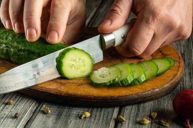 Elleri ustalıkla taze bir salatalığı tahta bir kesme tahtasıyla doğruyor. Mutfak ortamı sıcak ve davetkar. Yumuşak ışık sebzelerin canlı yeşilliğini güçlendiriyor..