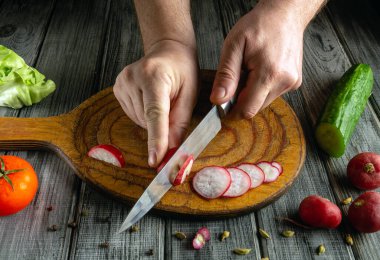 Eller ahşap bir kesim tahtasında parlak kırmızı turpları ustalıkla kesiyor. Etrafı marul, salatalık ve domates gibi taze sebzelerle çevrili..