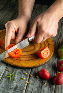 Kırsal ahşap bir kesme tahtasının üzerinde ustalıkla olgun domatesleri kesiyor. Tahtanın etrafında çeşitli taze sebzeler var. Sıcak bir mutfaktaki pişirme atmosferini arttırıyorlar..