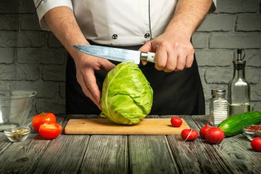 Bir şef ahşap kesme tahtasıyla marifetli bir şekilde lahana kesiyor. Şefin etrafında taze domatesler, salatalıklar, turplar ve canlı bir mutfak atmosferi sergileyen cam bir kâse bulunuyor..